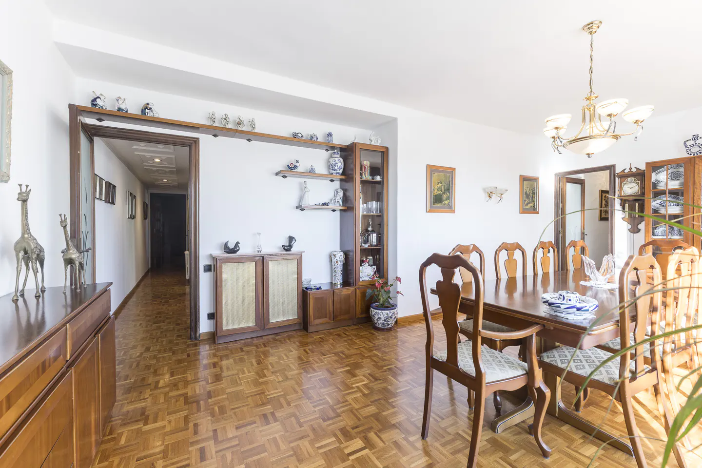 A bright dining room with a long wooden table and chairs, a chandelier, and parquet flooring. Shelves display decorative items.