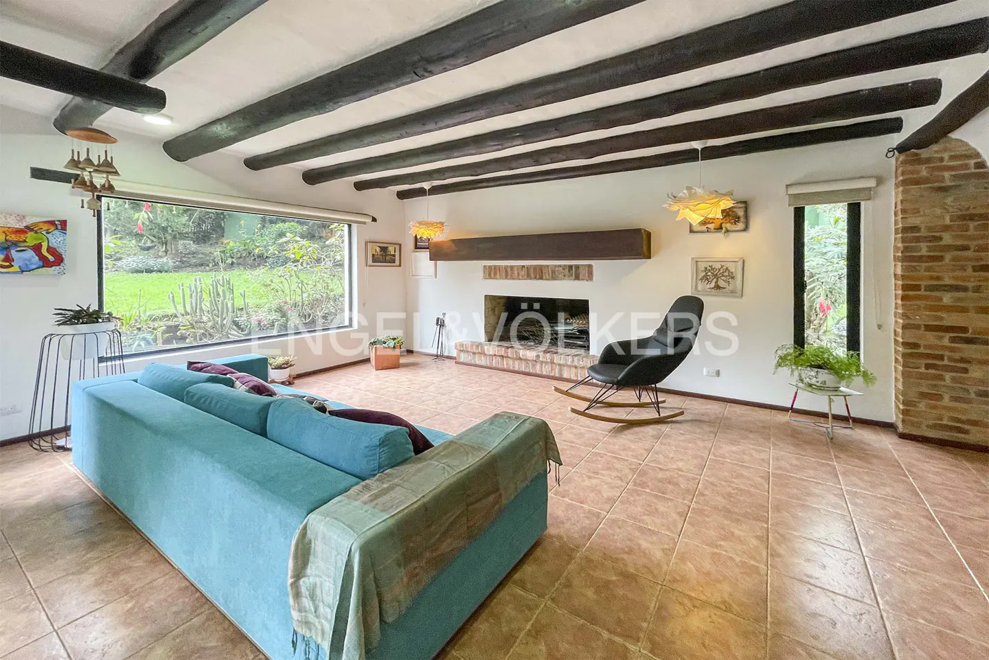 Living room with a turquoise sofa, black rocking chair, and a brick fireplace. Beamed ceiling and large window overlooking a green garden.