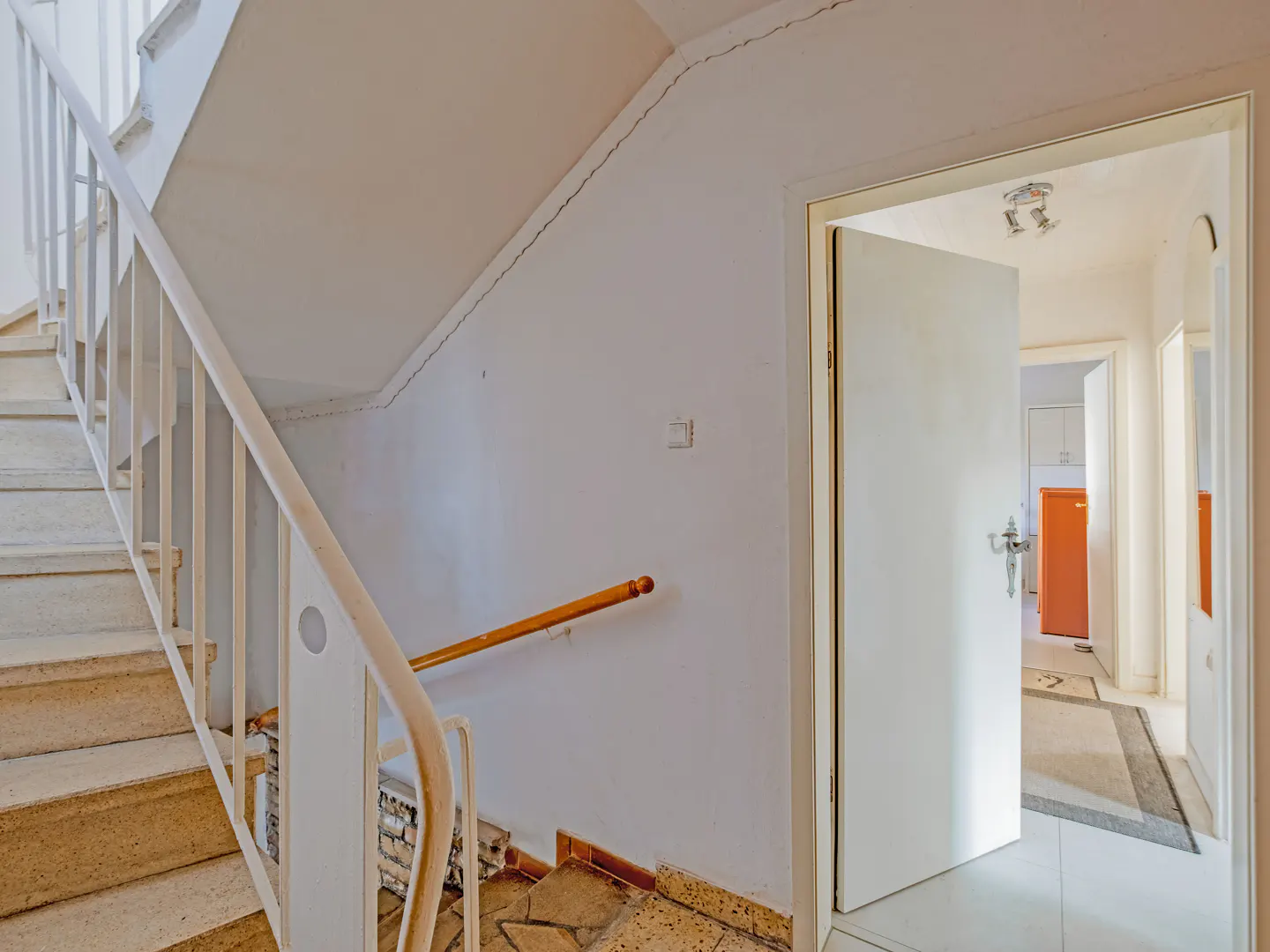 Interior shot of a stairwell with white walls, beige stairs and white railing. An open door reveals a hallway with an orange refrigerator.