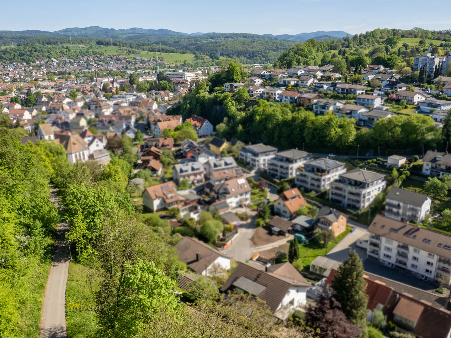 Aerial view of a town with houses, green trees, and mountains in the background on a sunny day.