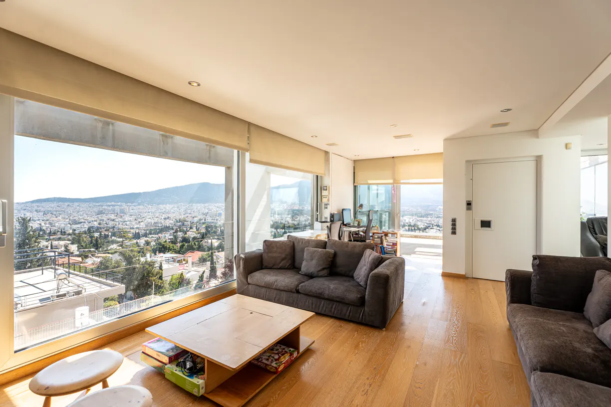 Bright living room with wood floors, gray sofas, and a light wood coffee table. Large windows offer a city view.