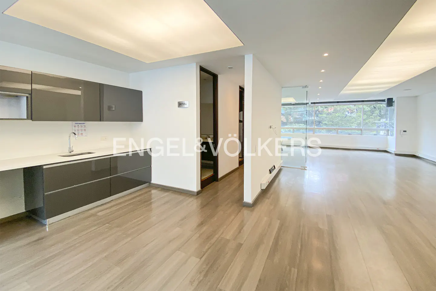 A bright, modern apartment interior with gray cabinets, light wood floors, and large windows overlooking greenery.