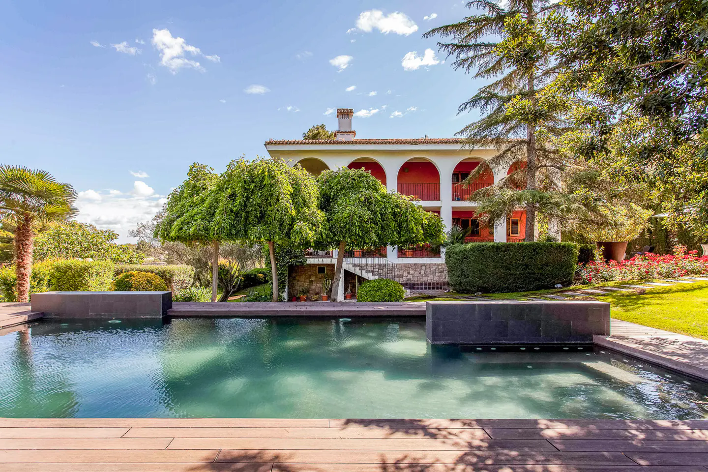 A large turquoise pool with a wood deck in front of a two-story white house with red accents and green trees.