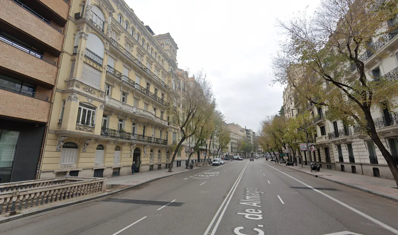 Street view of C. de Almagro in Madrid, Spain. Buildings line both sides of the street, with trees planted along the sidewalks. Cars are parked along the street.