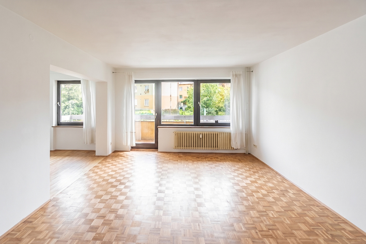 Bright, empty room with parquet floors, white walls, and large windows with white curtains. A radiator sits below the window.
