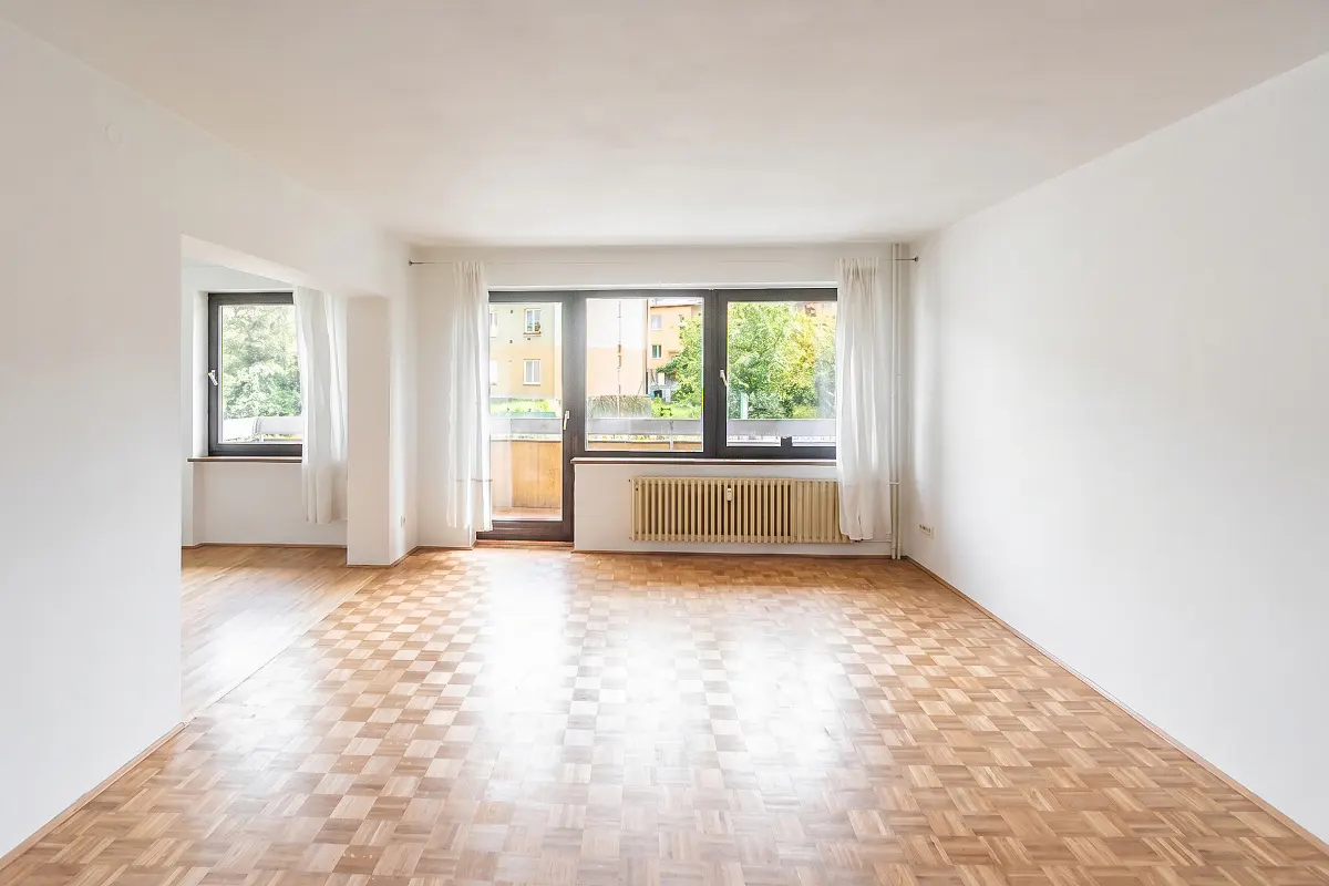 Bright, empty room with parquet floors, white walls, and large windows with white curtains. A radiator sits below the window.