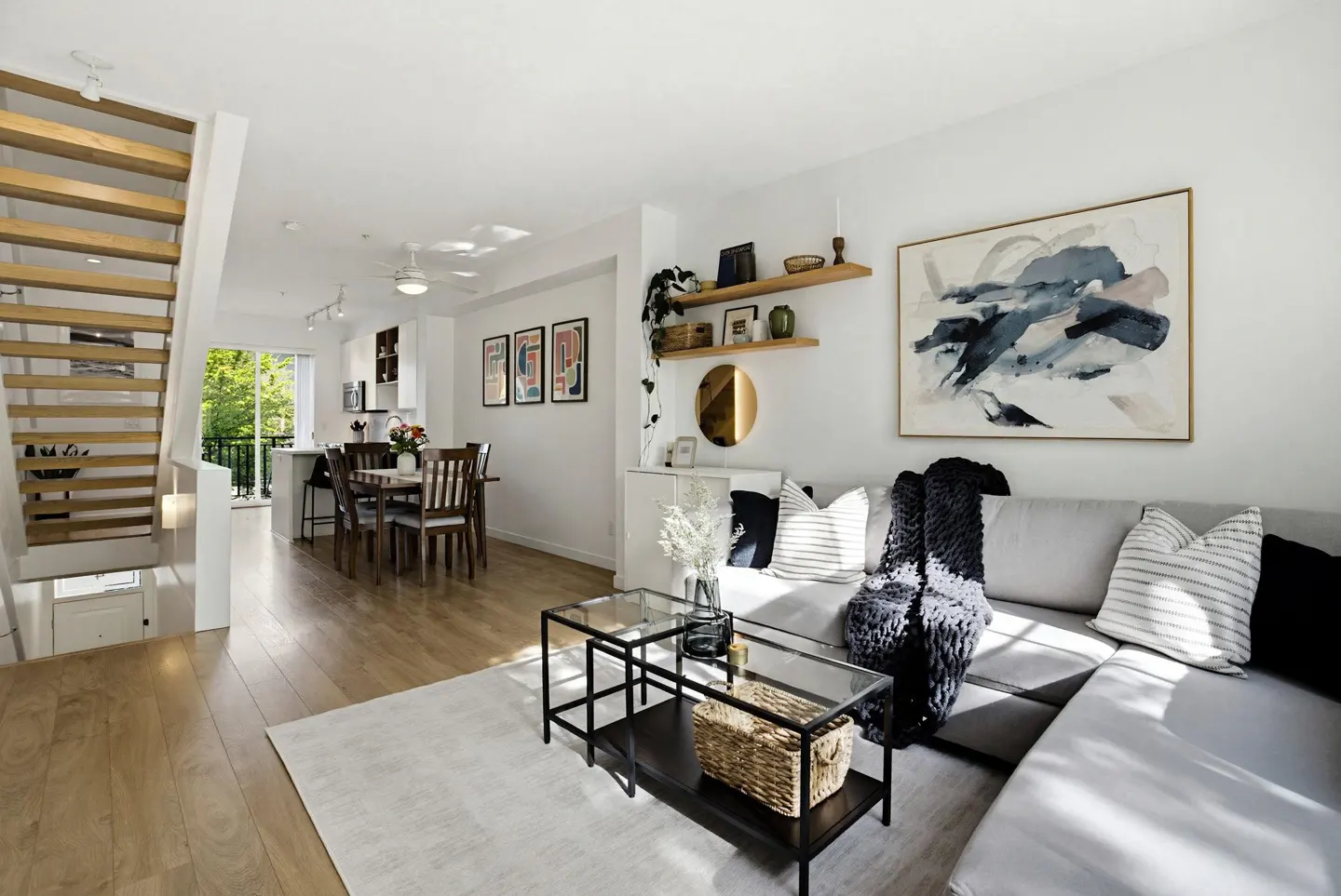 Bright, open-concept living space with wood floors, white walls, and a gray sectional sofa. Stairs, dining area, and kitchen are visible.