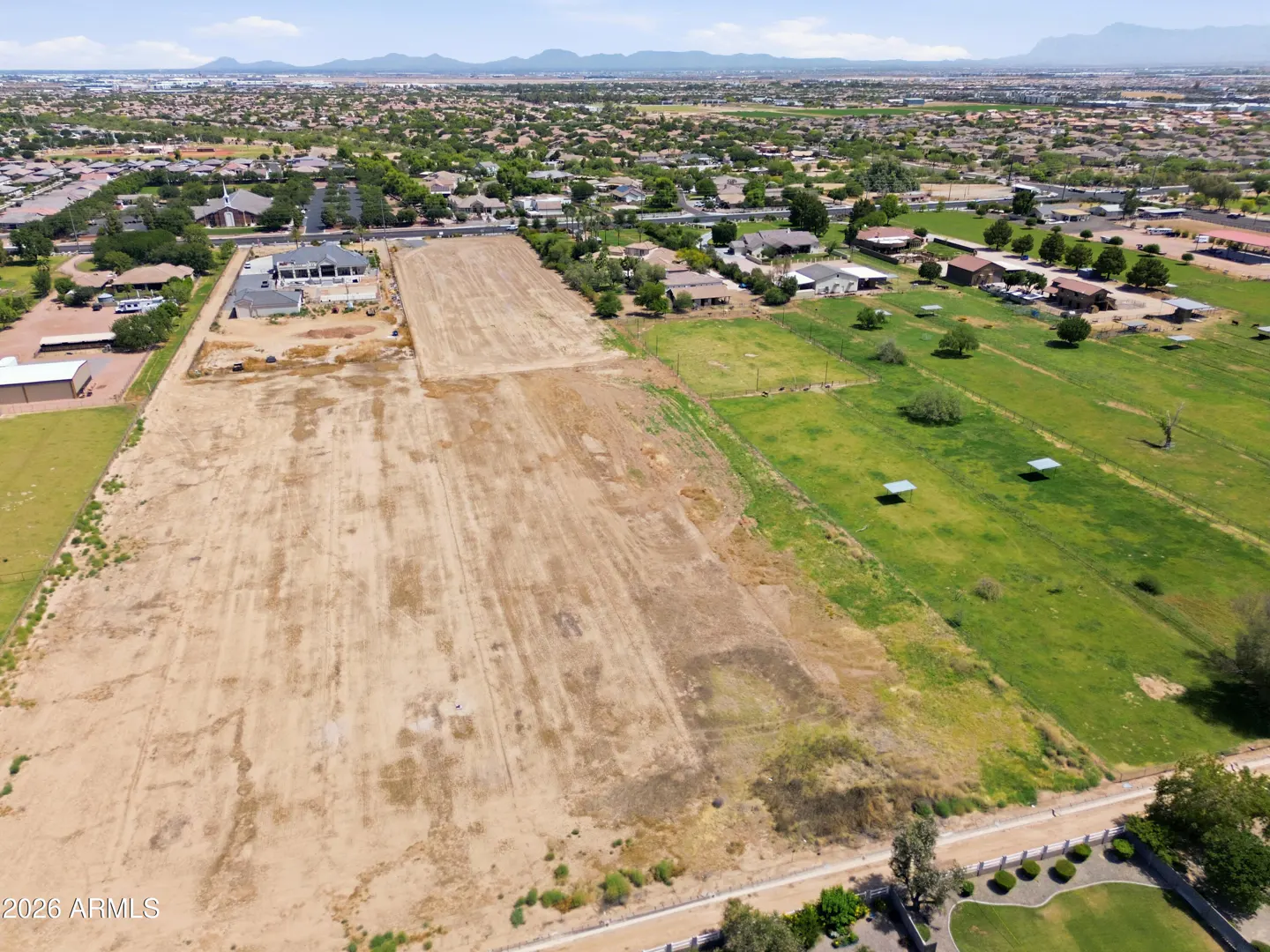 Aerial view of a large, rectangular vacant dirt lot bordered by green fields and suburban homes under a clear blue sky.
