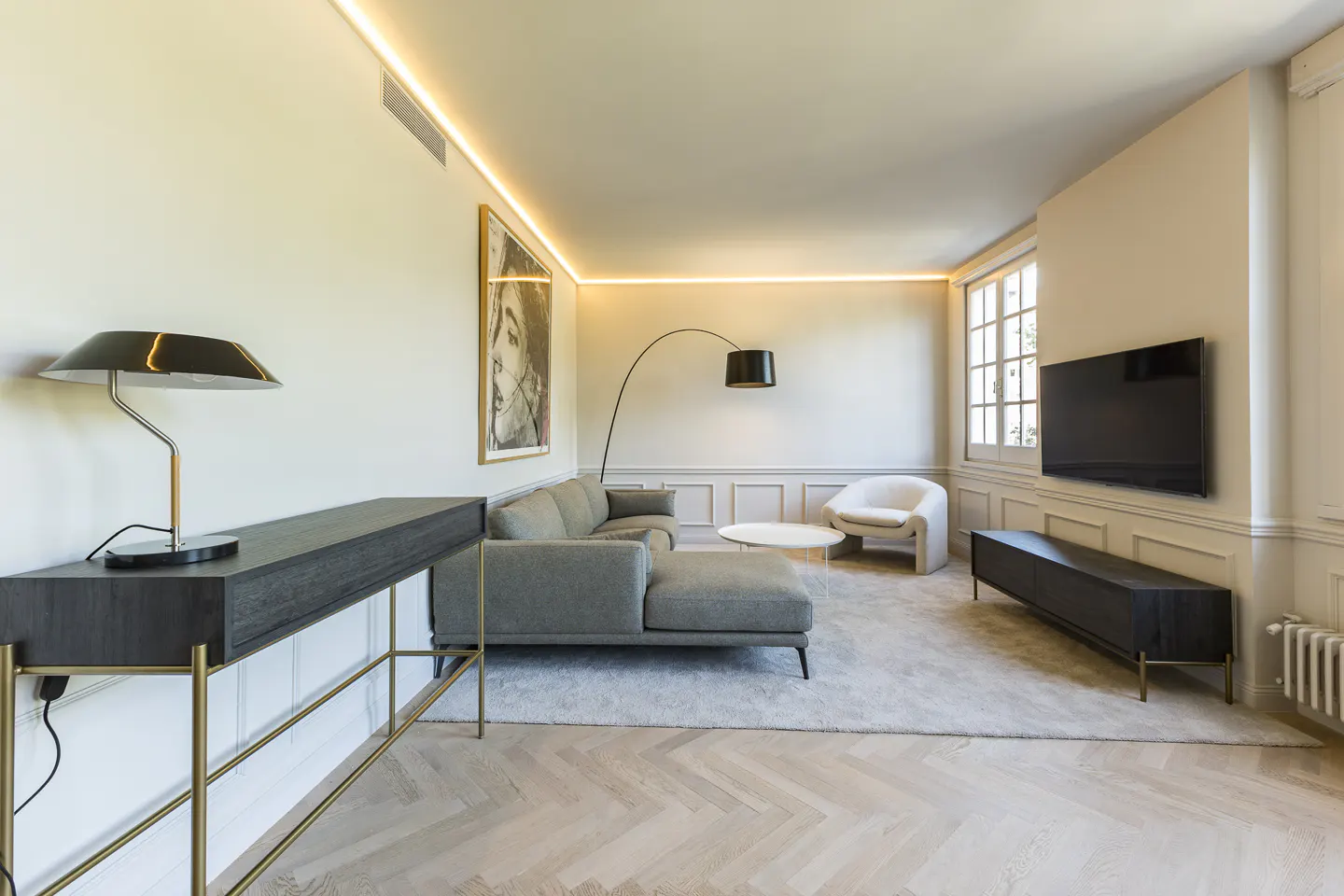 A bright living room with a gray sofa, white chair, and black console table. A large TV hangs on the wall near a window. The floor is light wood in a herringbone pattern.