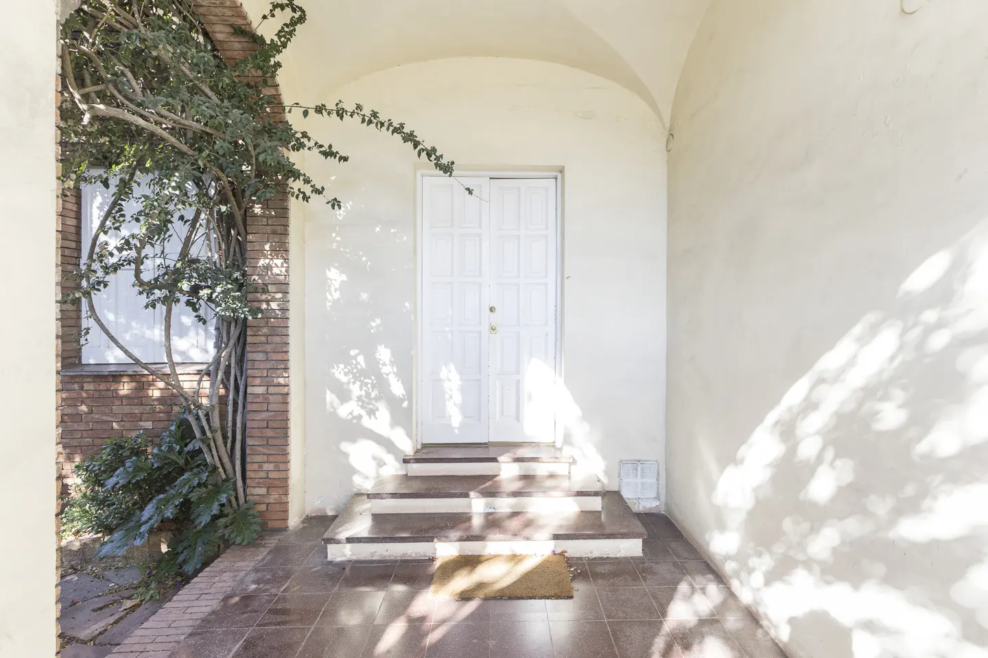 Exterior view of a home's entrance with a white door, steps, tile floor, and brick wall with climbing vines.
