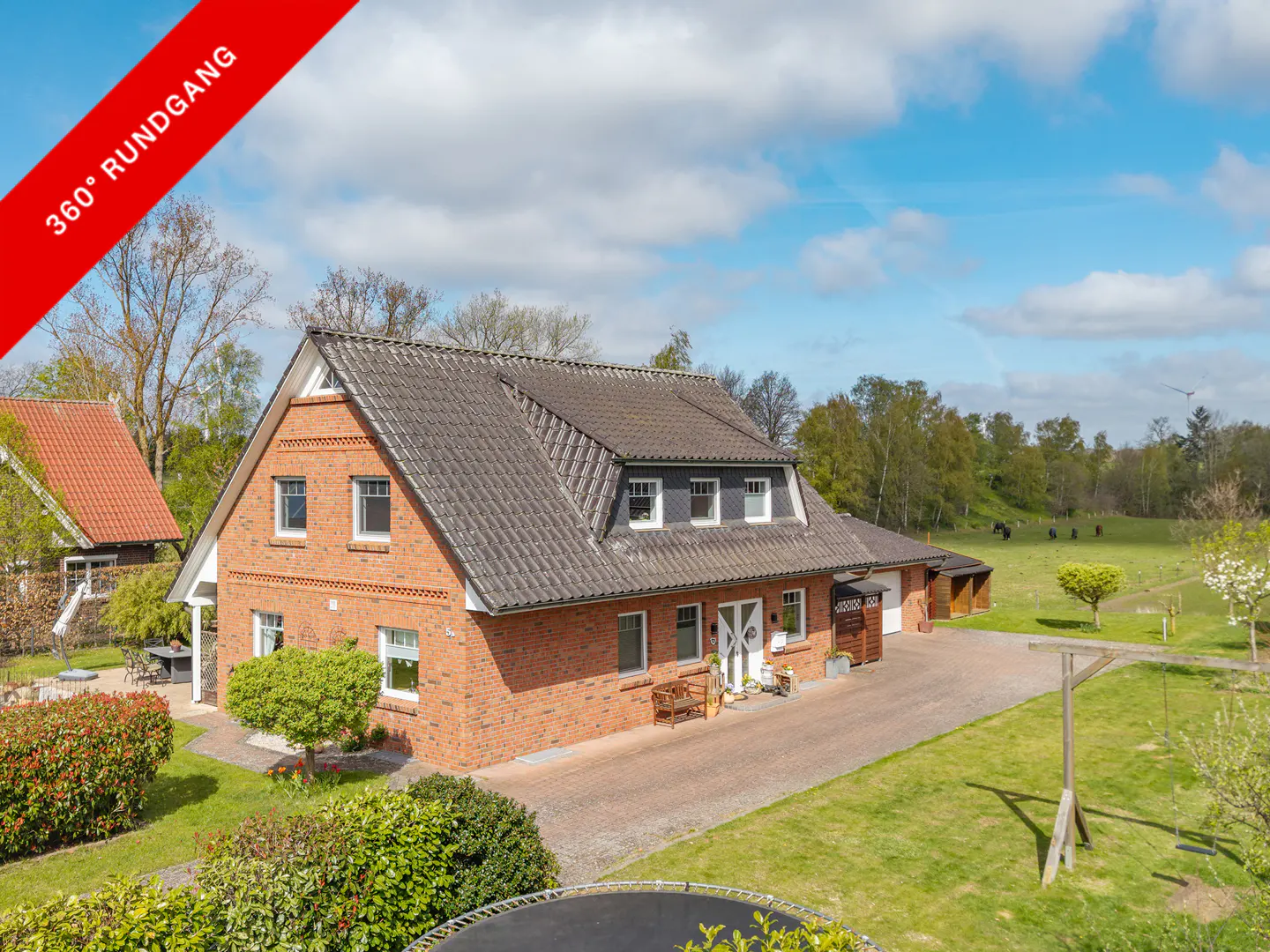 Two-story brick house with a gray roof, surrounded by green lawn, trees, and a field with grazing animals under a blue sky.