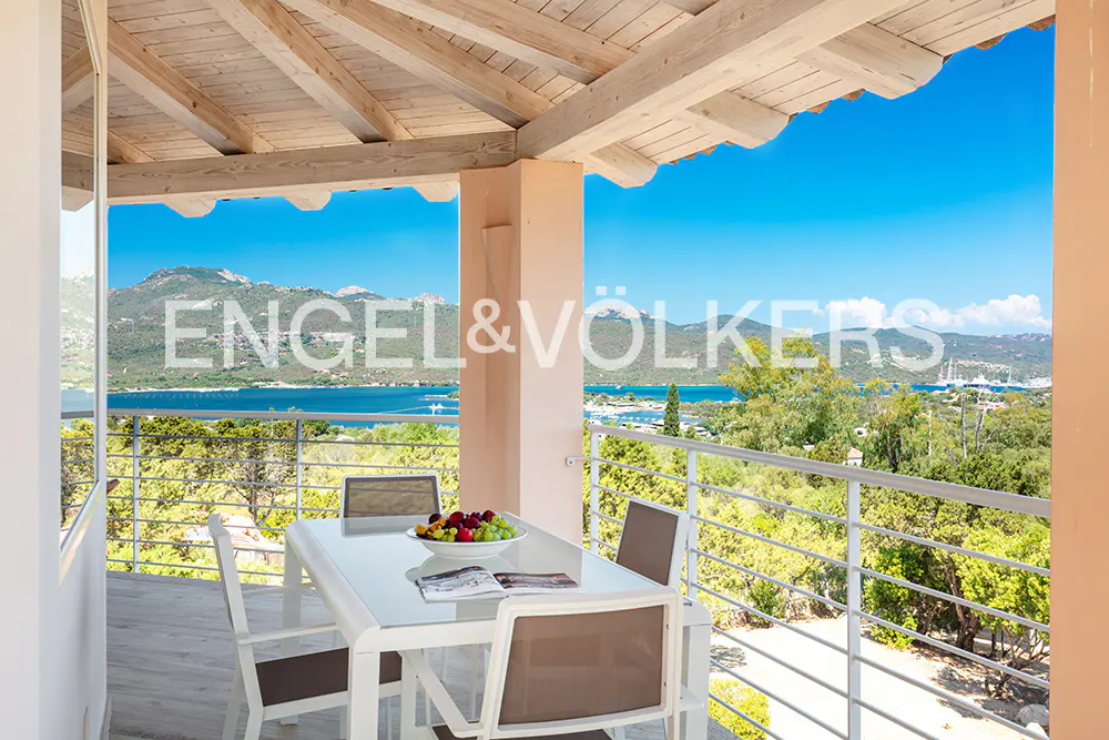 Covered balcony with a dining table, fruit bowl, and chairs overlooking a scenic bay with mountains under a blue sky.