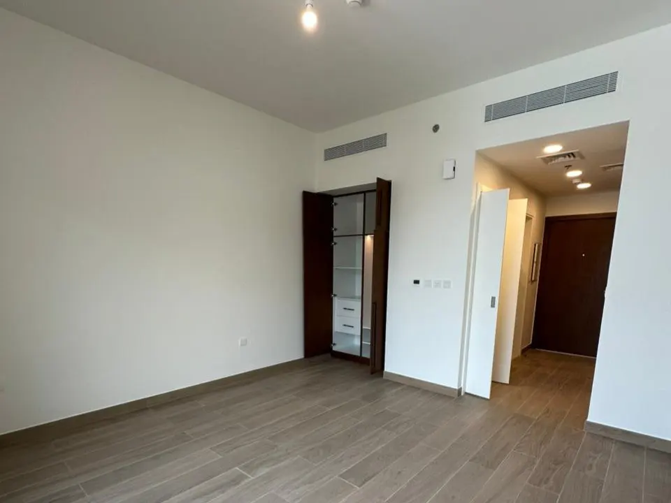 A bright, empty room with wood-look tile floors, white walls, and a dark wood cabinet with open doors.