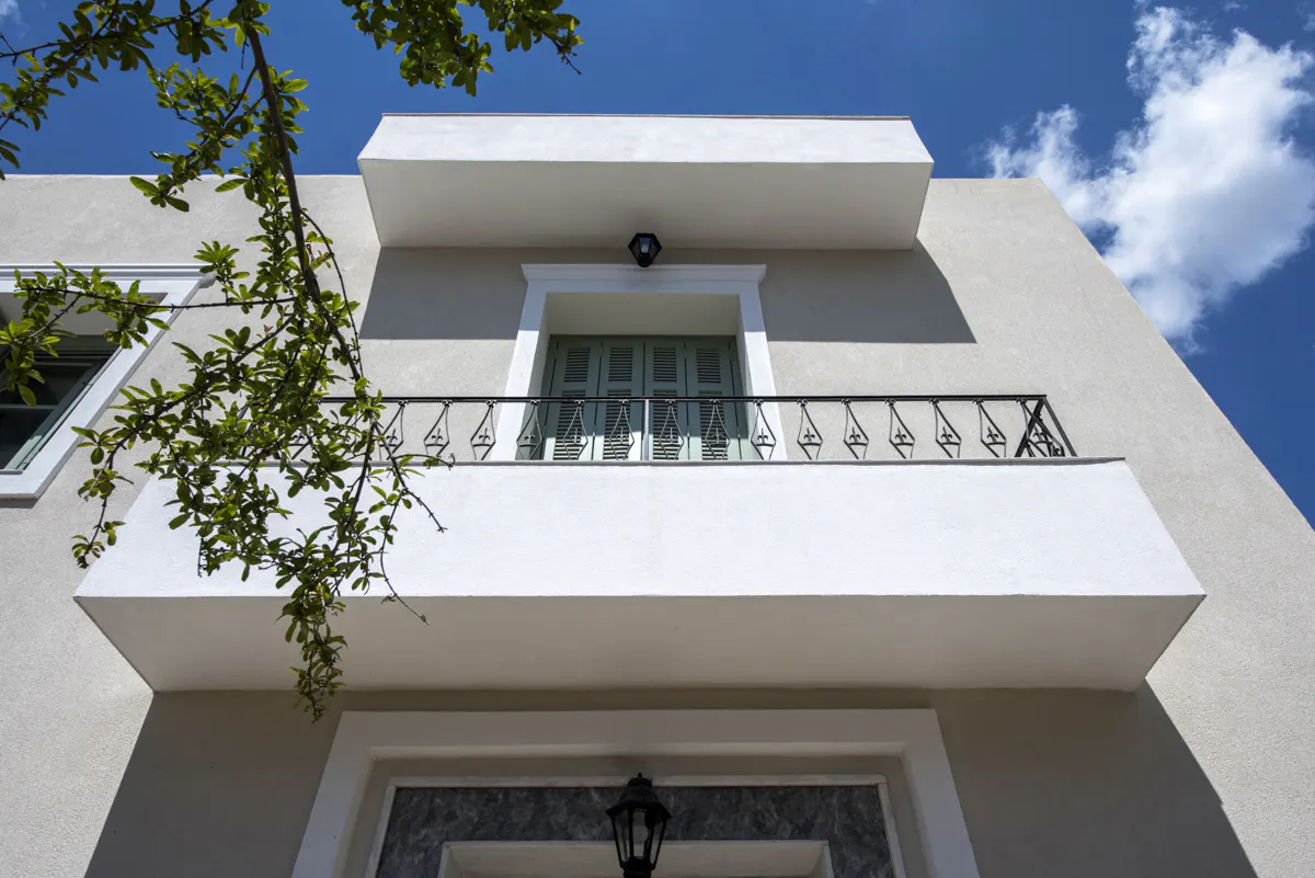 Exterior view of a two-story building with a balcony, light-grey walls, white trim, and a blue sky background.