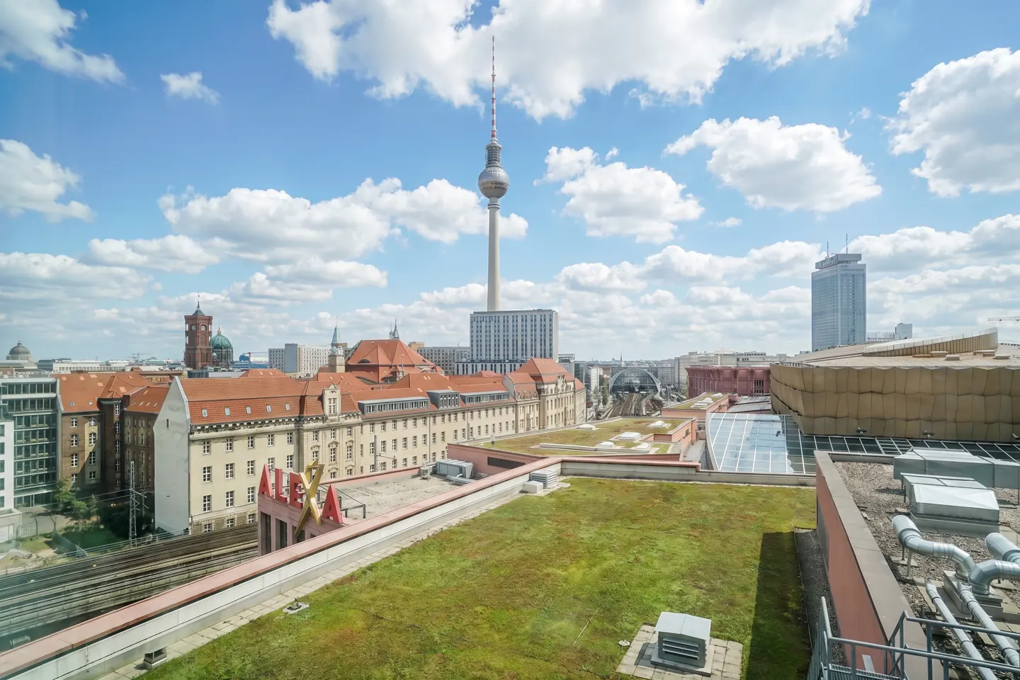 Berlin cityscape view with the TV Tower, red-roofed buildings, green roofs, and a blue sky with white clouds.