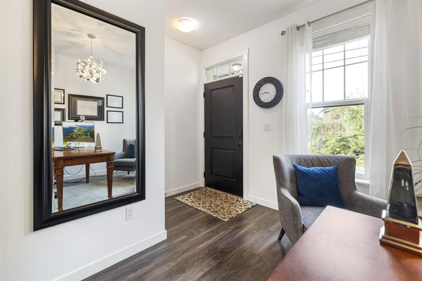 Entryway with dark wood floors, white walls, and a black front door. A large mirror reflects a desk and chair. A gray chair sits near a window with white curtains.