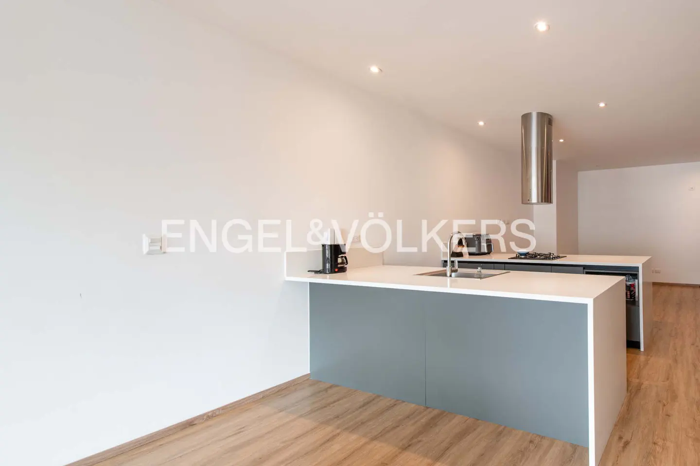 Modern kitchen with gray and white island, stainless steel appliances, and wood floors. Engel & Völkers logo on the wall.