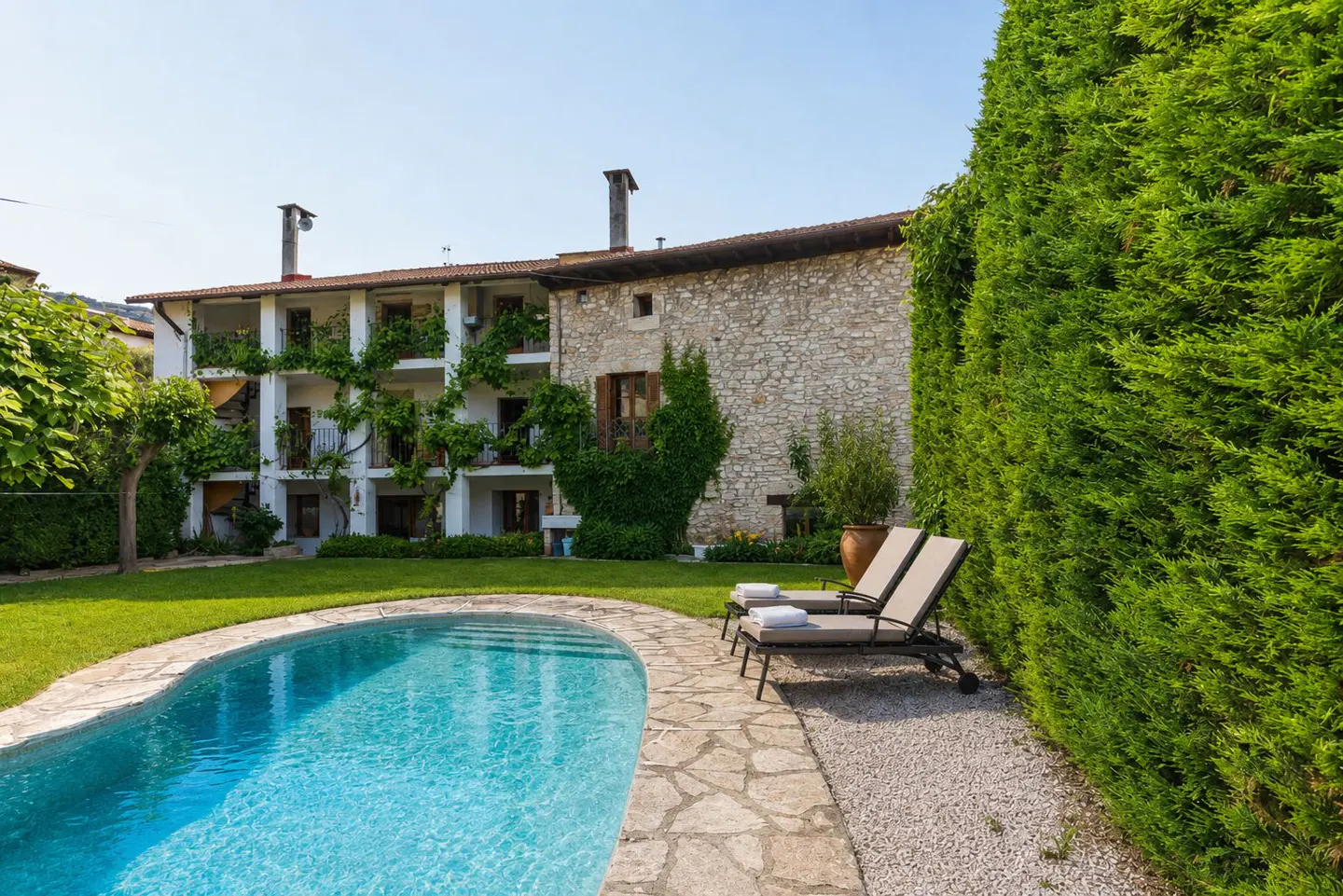 Exterior view of a stone house with a pool, lawn, and two lounge chairs.