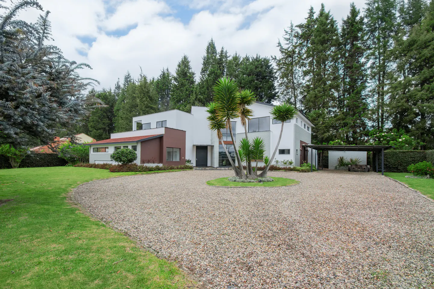 Modern white house with brown accents, a gravel driveway, and palm trees in the center. Tall green trees surround the property.