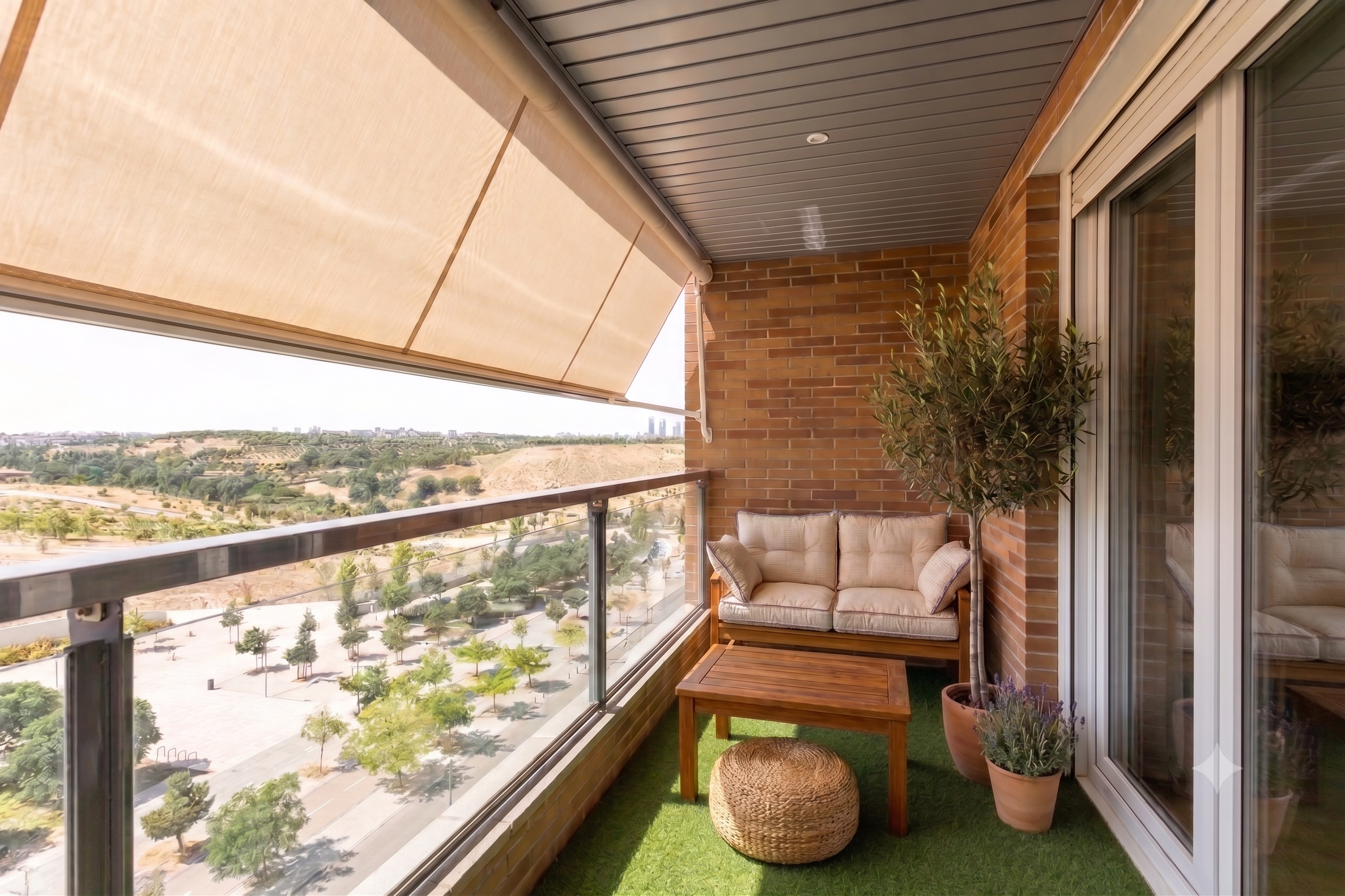 A balcony with a beige awning, brick wall, wooden sofa, coffee table, pouf, potted plants, and a view of a park.