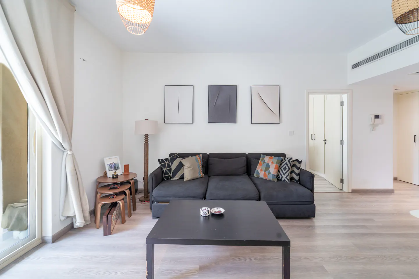 Living room with a gray sofa, black coffee table, and three framed art pieces on a white wall. A stack of wooden tables sits near a window with beige curtains.