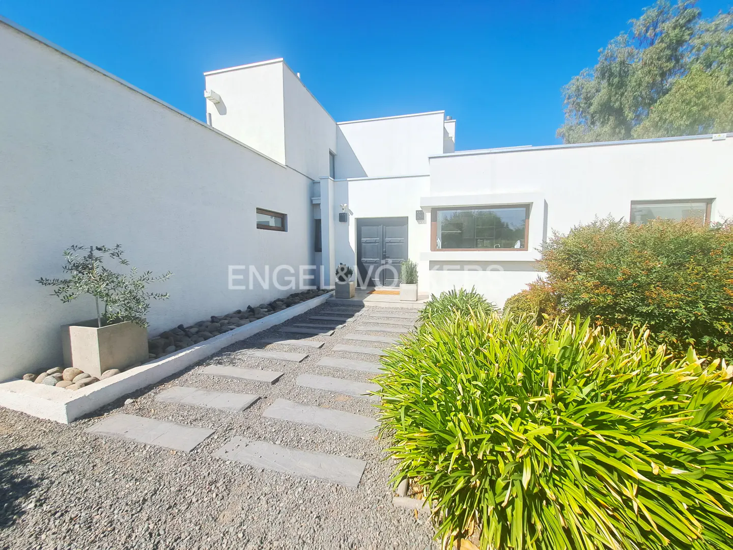 Modern white house with a stone path leading to the front door, surrounded by green bushes and a blue sky.
