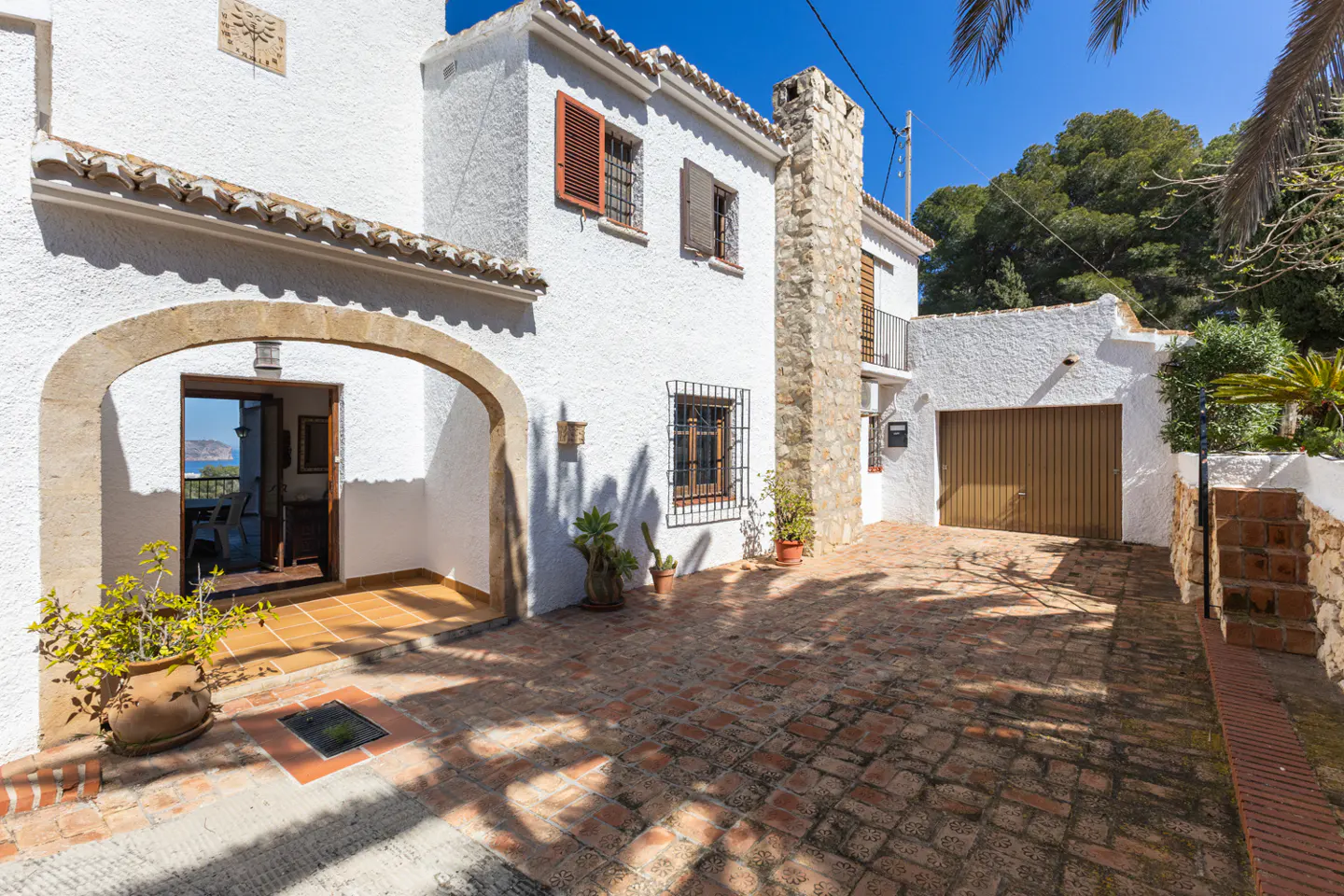 Exterior view of a white stucco house with a stone chimney and brick driveway. An arched entrance leads into the home.