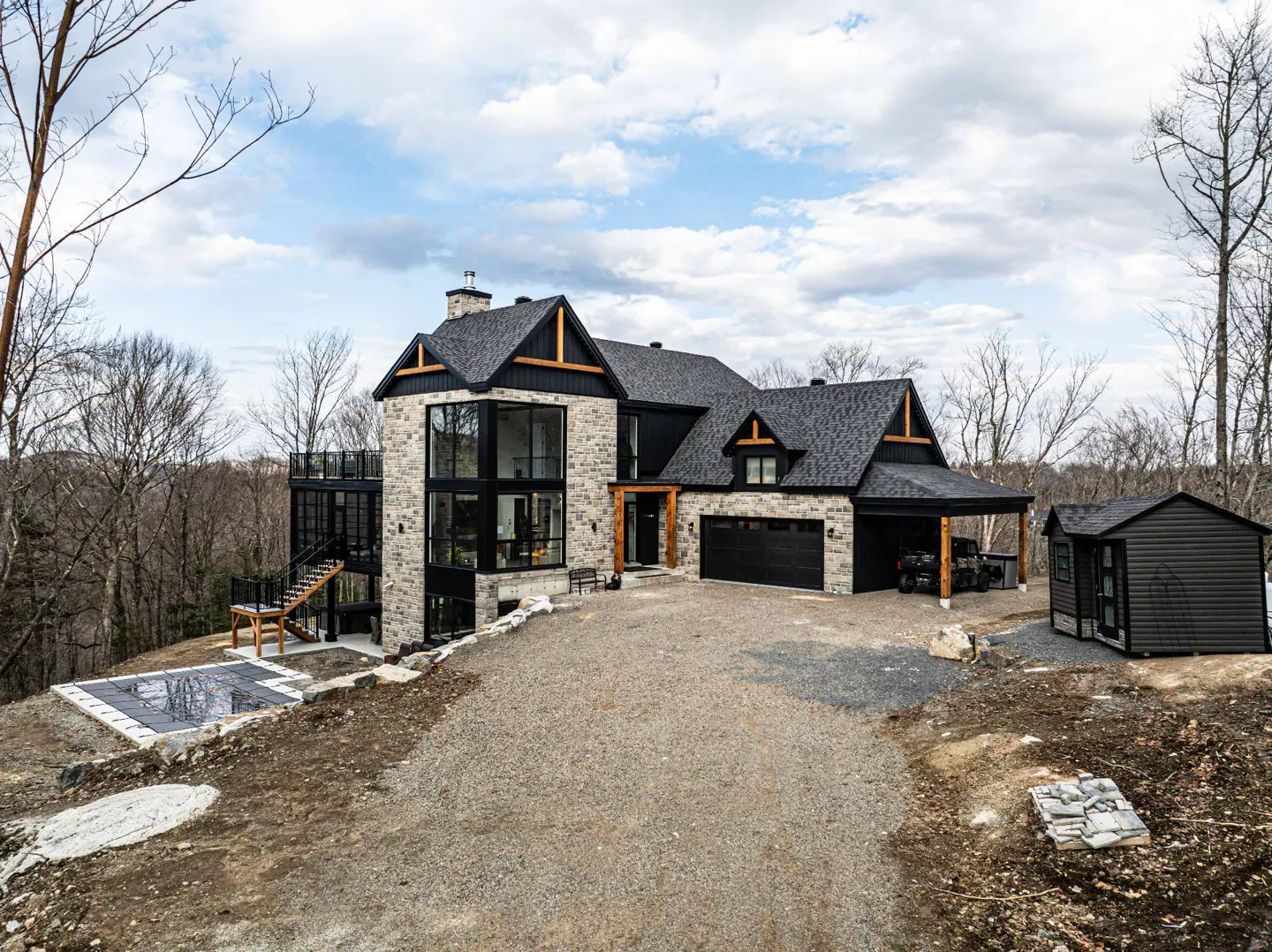 Exterior of a modern stone house with black trim, a black roof, and a gravel driveway. A pool and shed are visible.