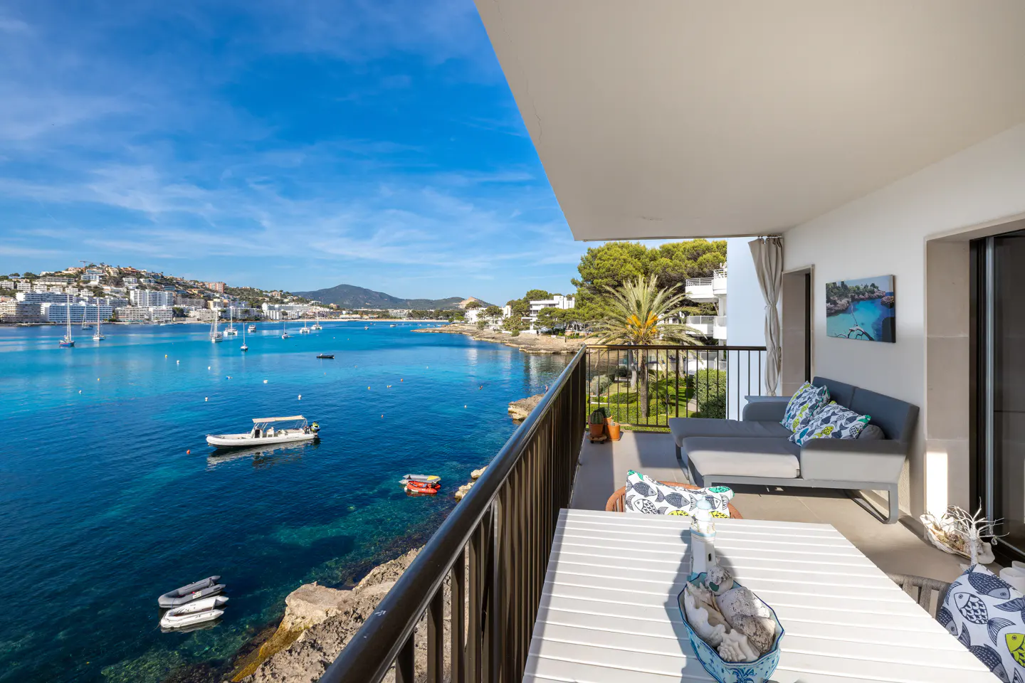 Balcony view of blue ocean with boats. Table and gray sofa with patterned pillows on the patio. Sunny day.