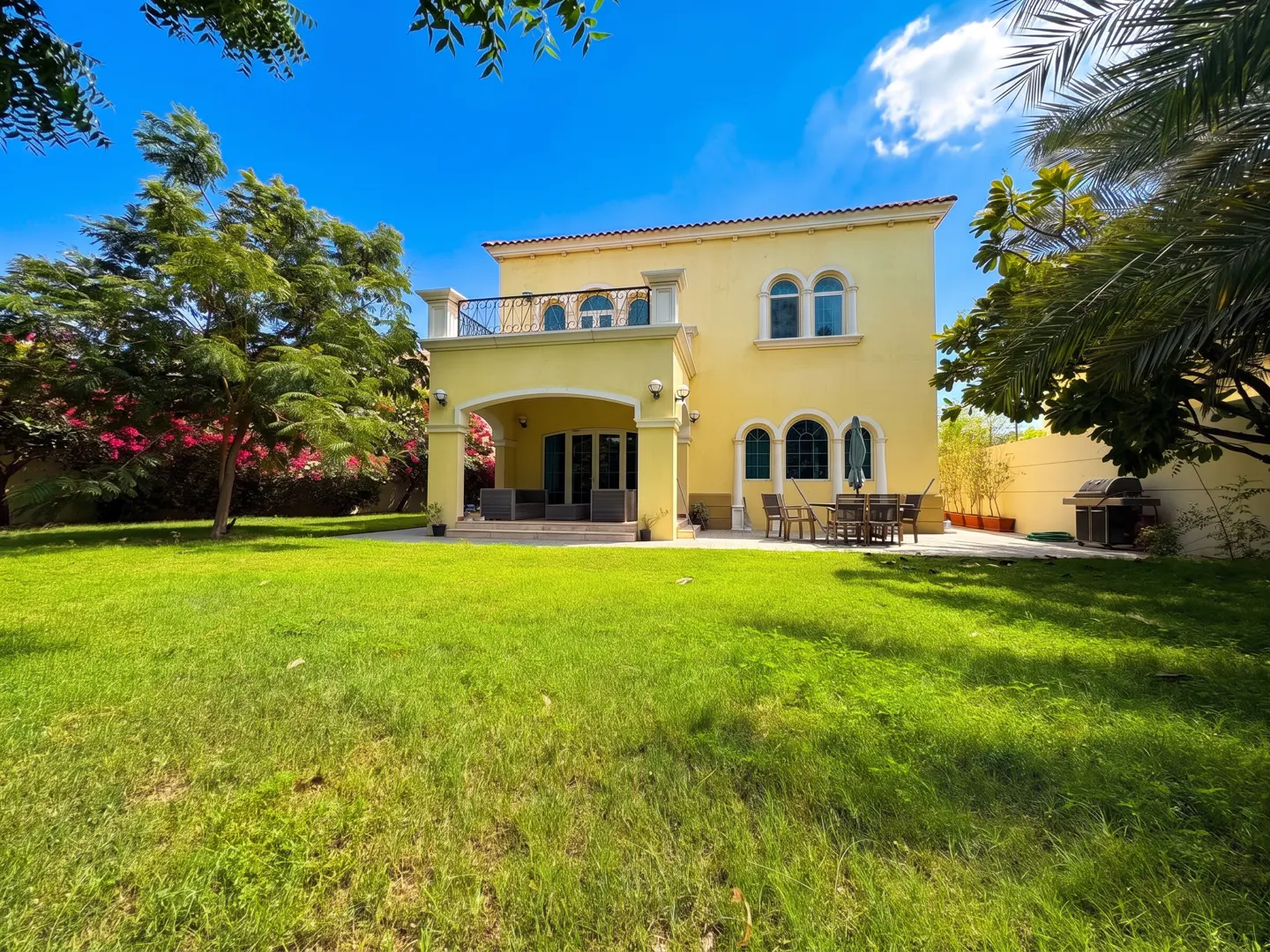 Yellow two-story villa with a patio, outdoor dining set, and a large green lawn under a bright blue sky.
