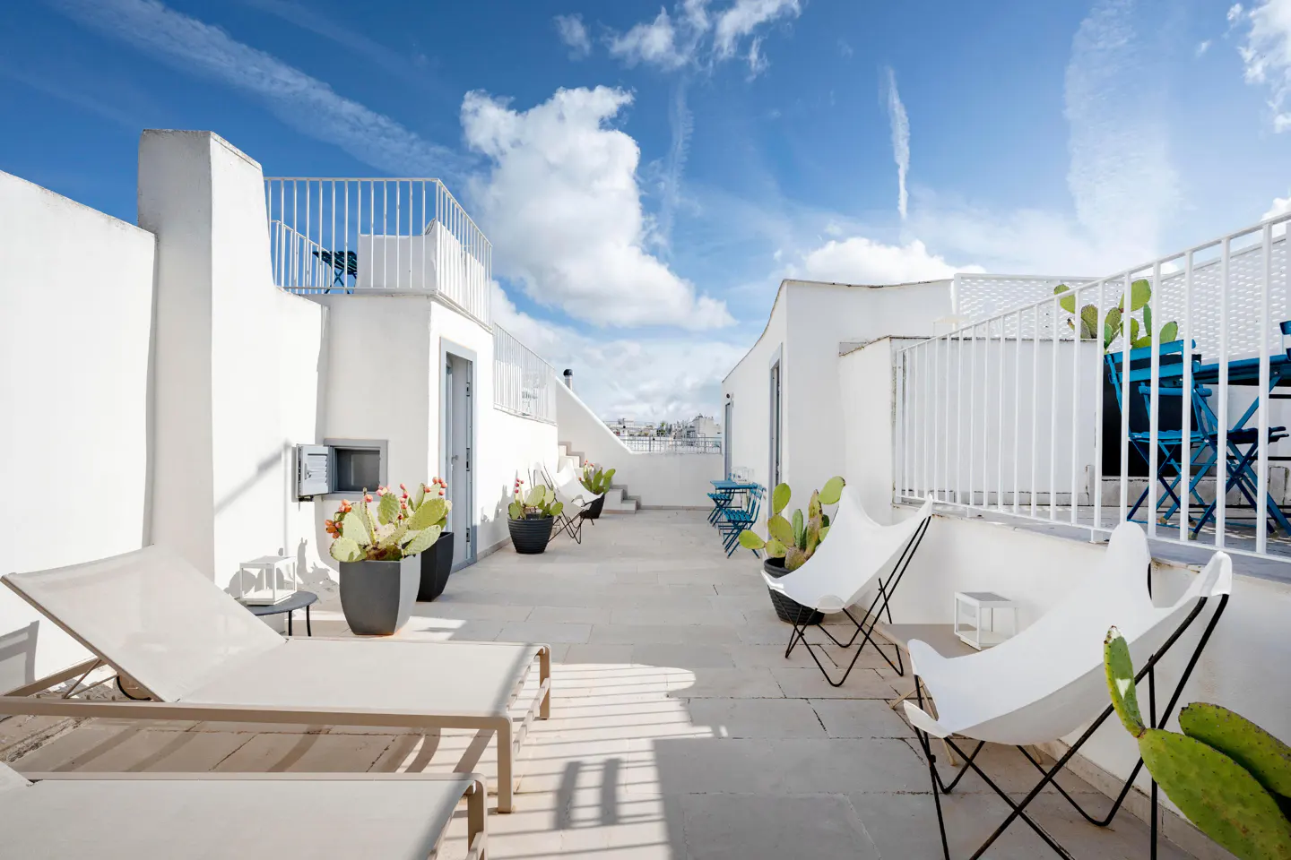 Rooftop patio with white walls, lounge chairs, and potted cacti under a blue sky with clouds.