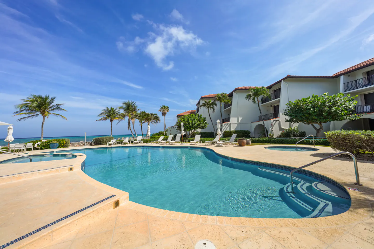 Outdoor pool with turquoise water, surrounded by palm trees and white lounge chairs, next to a white building with balconies under a blue sky.