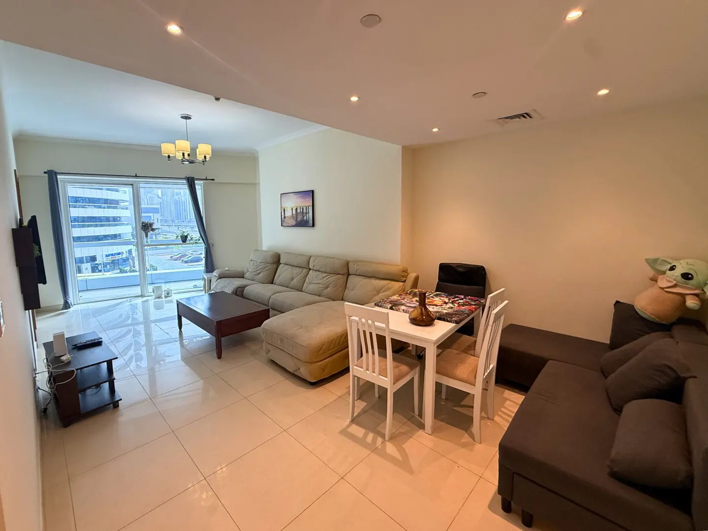 A bright living room with beige walls, tile floors, and a large window with city views. A beige sectional sofa, a dark brown sofa, and a white dining table with chairs.