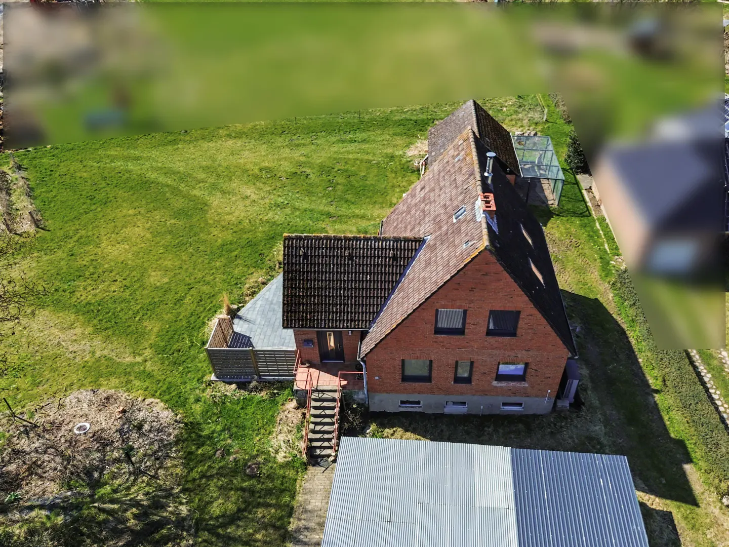 Aerial view of a red brick house with a dark roof, surrounded by a green lawn and a metal shed.