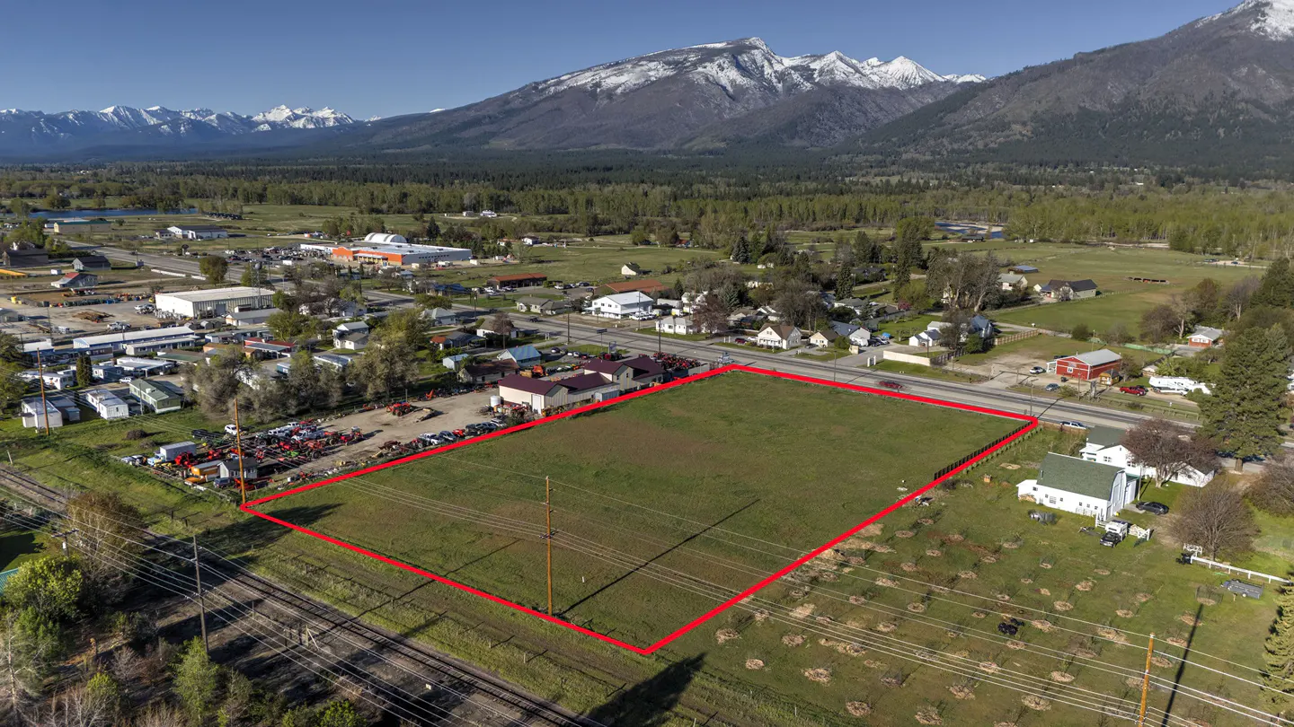 Aerial view of a grassy lot outlined in red, with mountains, trees, and buildings in the background.