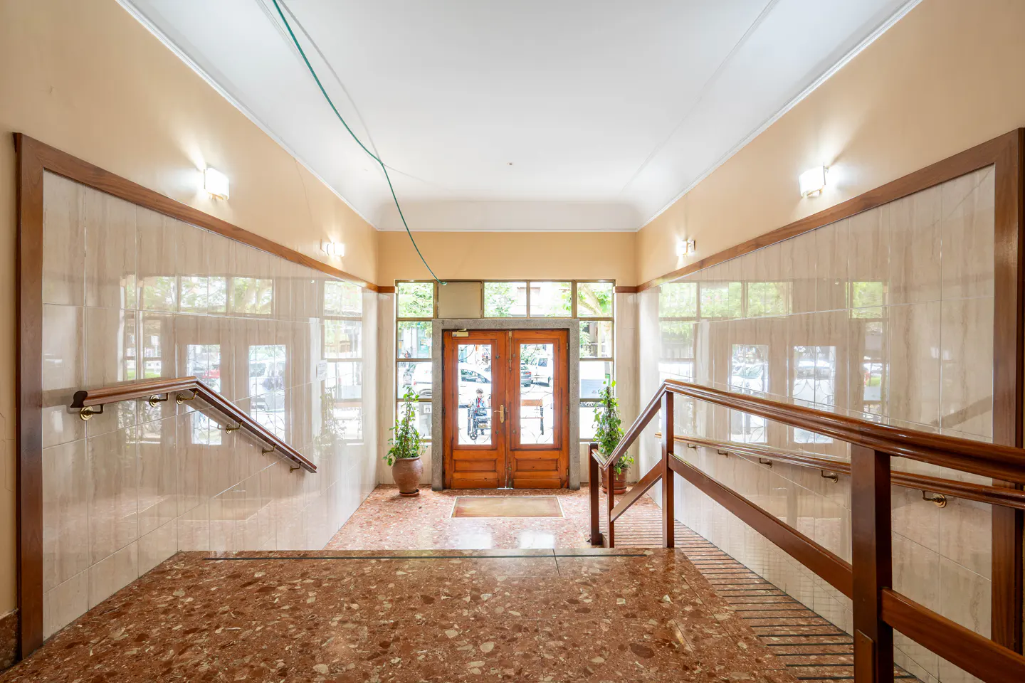 Interior view of a building entrance with marble floors, tiled walls, and wooden handrails leading to double doors.