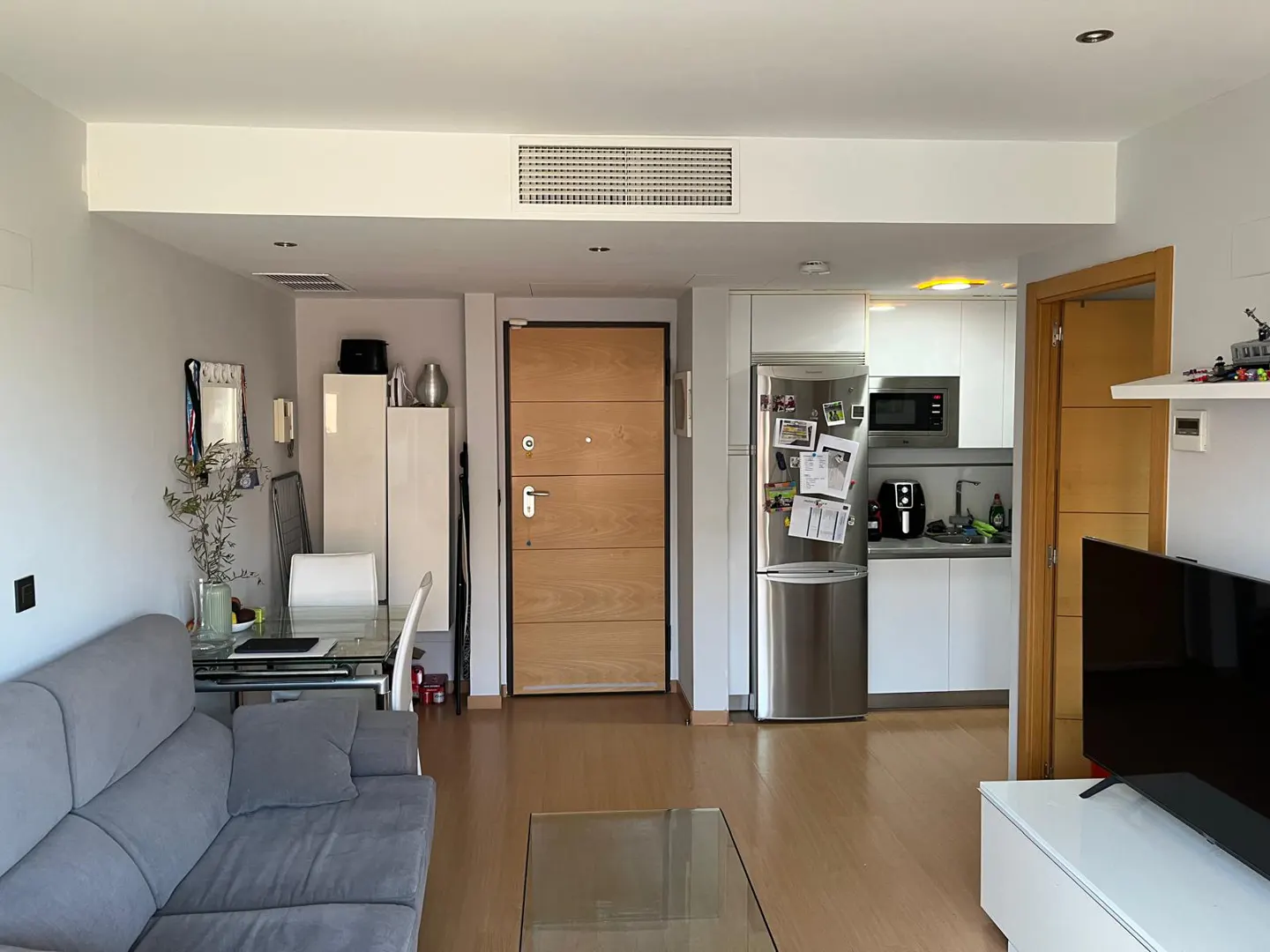 A bright apartment interior with a gray sofa, glass table, and a modern kitchen with stainless steel appliances.