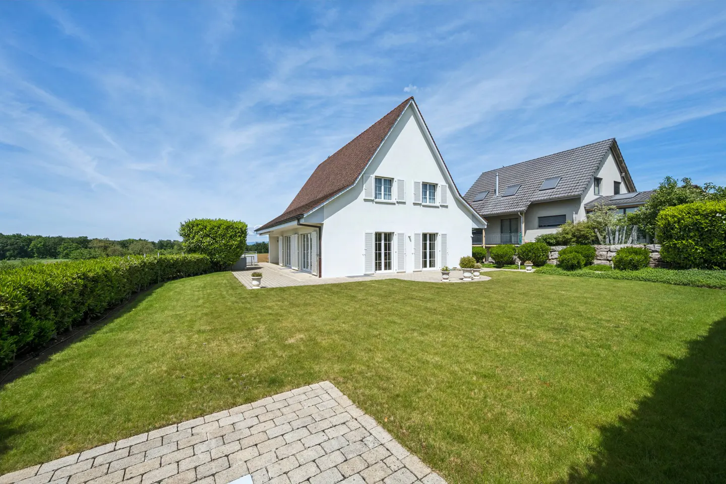 A white house with a brown roof sits on a green lawn under a blue sky. Another house is visible in the background.