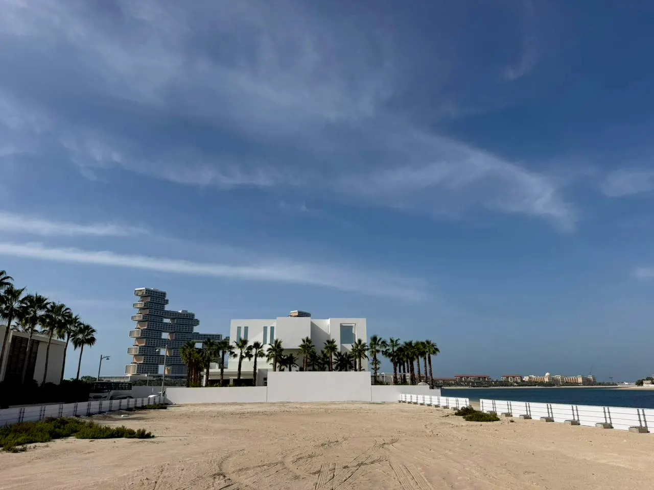 A vacant sandy lot with palm trees, modern white buildings, and a blue sky with wispy clouds in the background.