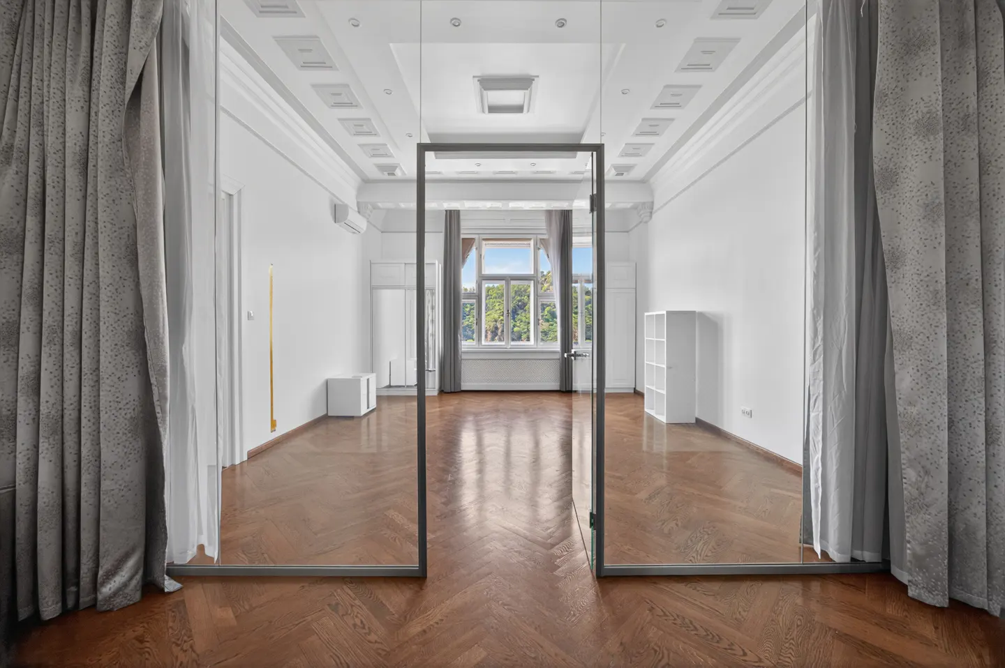 A bright, empty room with herringbone wood floors, white walls, and a glass door. Gray curtains frame the view.