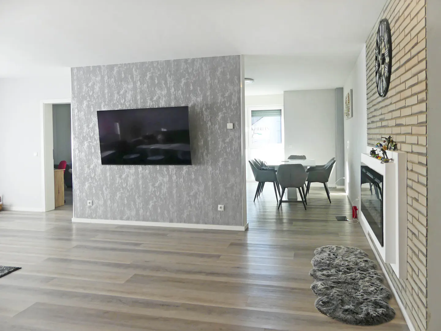 Open-concept living and dining area with gray wood floors. A TV is mounted on a silver accent wall. Dining table with gray chairs is visible.