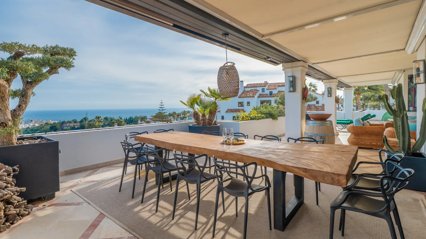 Outdoor dining area with a long wooden table, black chairs, and ocean view. A woven pendant light hangs above the table.