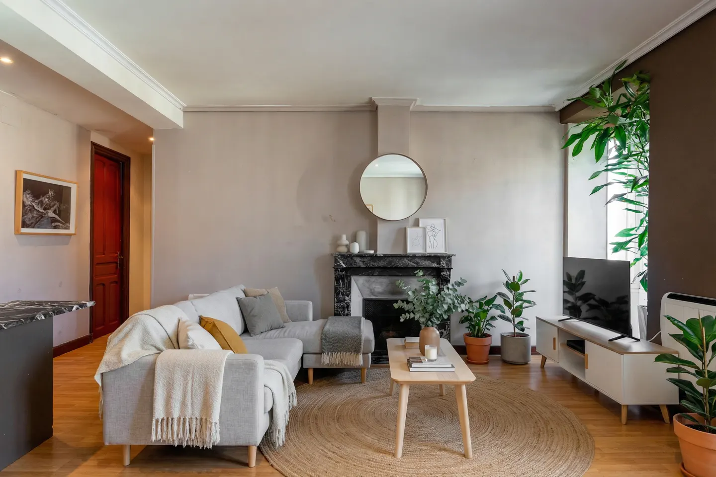 Living room with a gray sectional sofa, round jute rug, marble fireplace, and houseplants. A round mirror hangs above the fireplace.