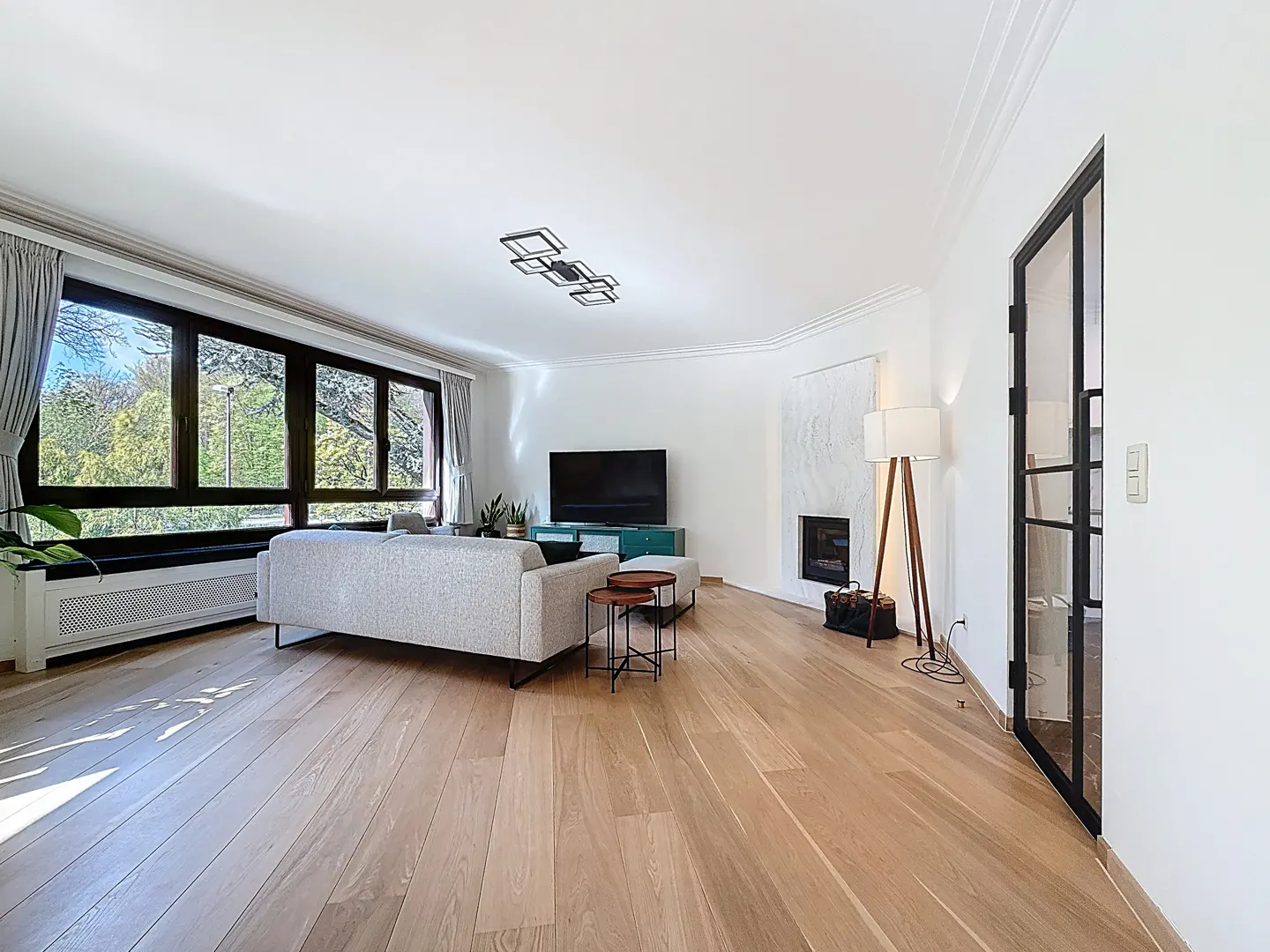 Bright living room with wood floors, a gray sofa, a black TV, and a modern fireplace. Large windows offer a view of trees.