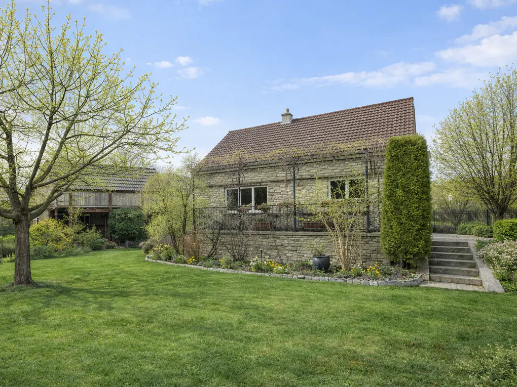 Exterior view of a stone house with a brown roof, green lawn, and trees under a blue sky.