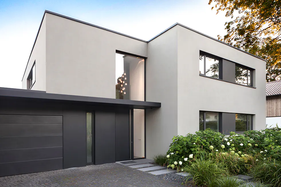 Modern two-story home with a gray exterior, black trim, and a flat roof. A large window shows a chandelier inside. Green landscaping in front.