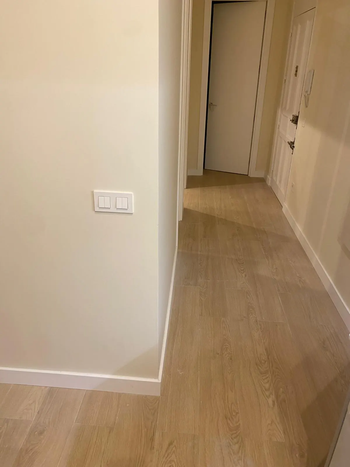 Hallway with light wood-look tile floor, cream walls, white trim, and white light switches. Doors are visible in the background.