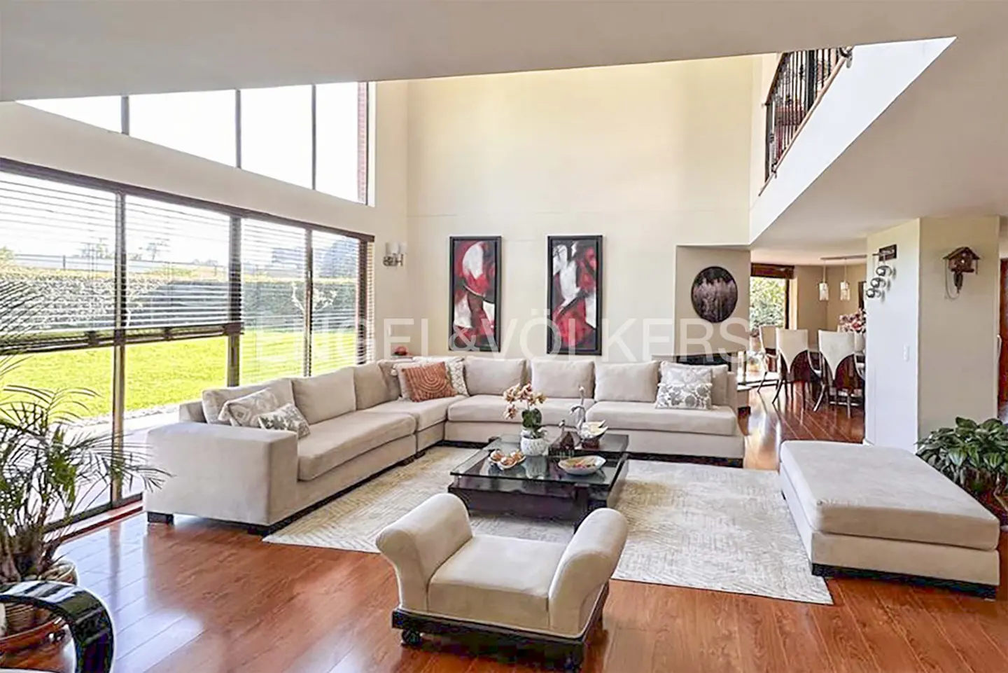Bright living room with a beige sectional sofa, glass coffee table, and hardwood floors. Large windows offer a view of the green lawn.