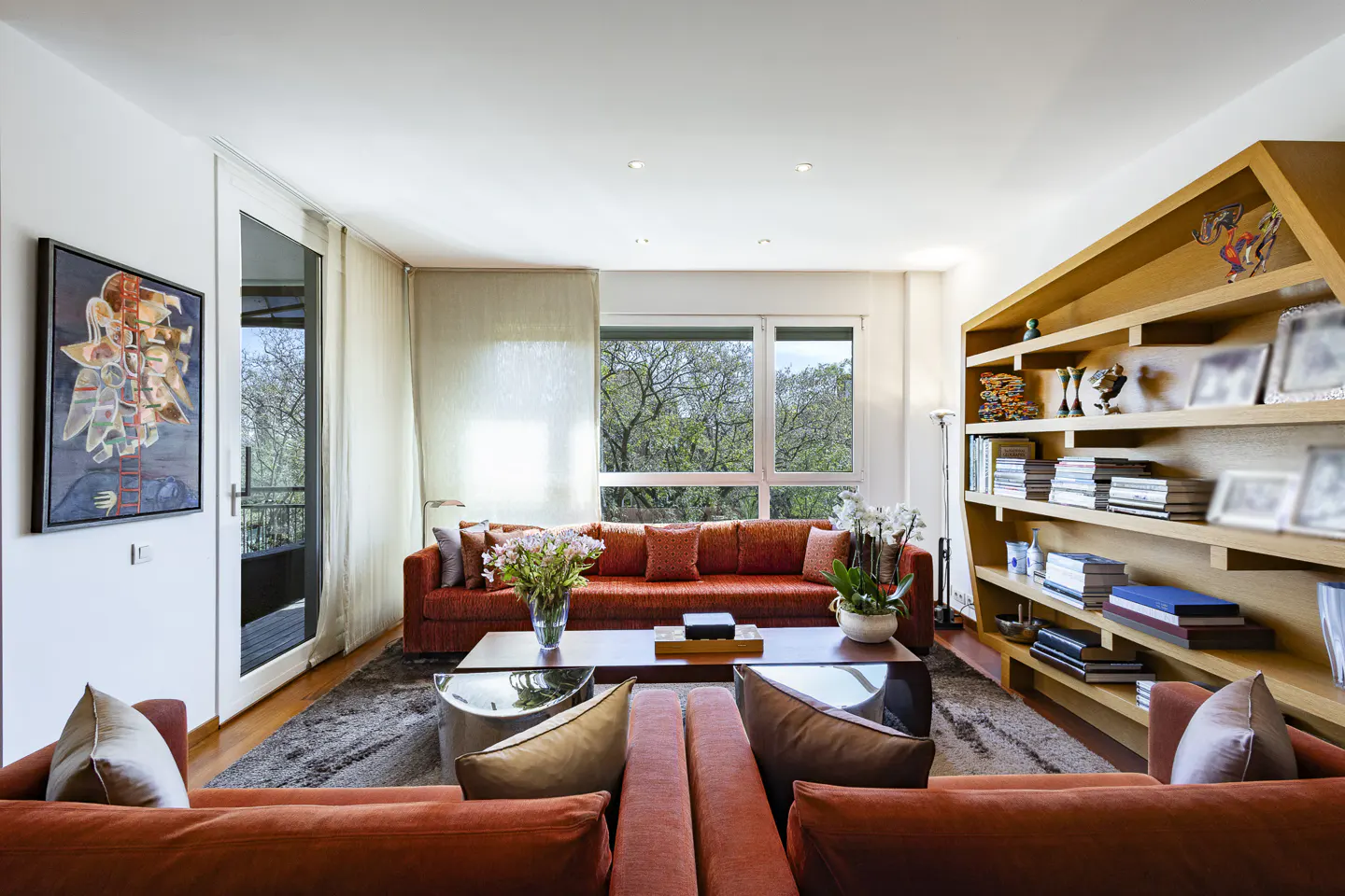 Bright living room with orange sofas, a wooden bookshelf, and a large window overlooking trees. A modern art piece hangs on the wall.