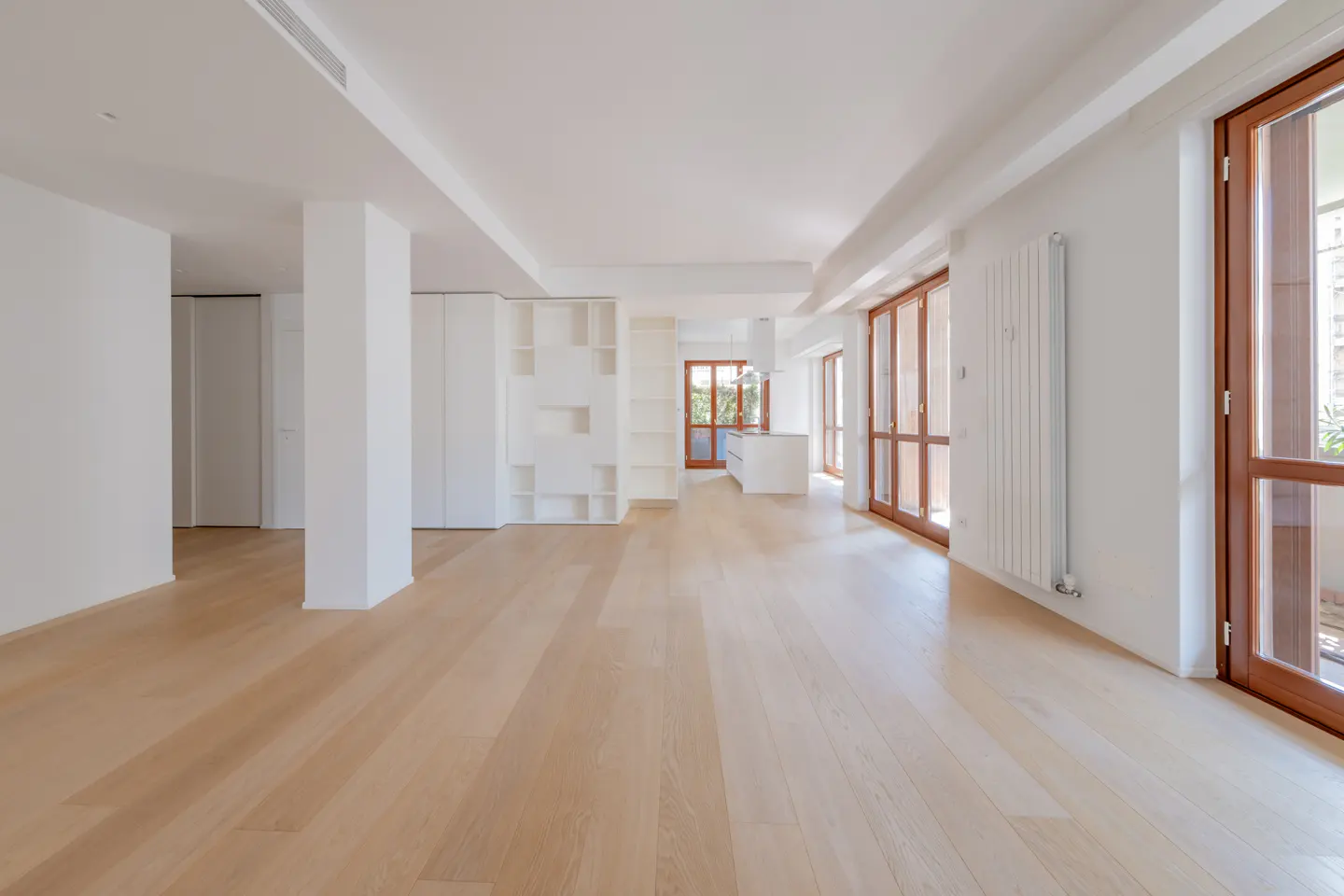 Bright, empty room with light wood floors, white walls, and brown wood-framed windows. A white shelving unit and kitchen are visible in the background.